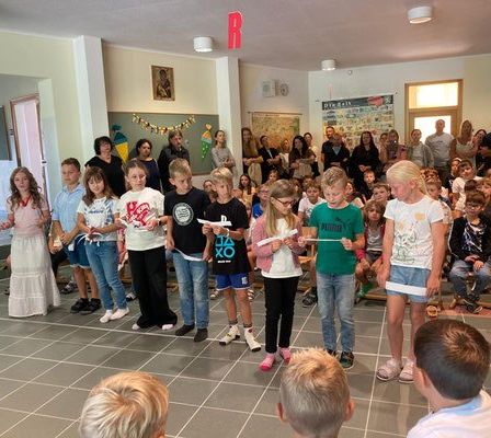 Eine Gruppe von Kindern steht im Kreis, Papiere in der Hand, vielleicht im Proben für ein Schulstück, während Erwachsene und Kinder ihnen in einem Raum zuschauen.