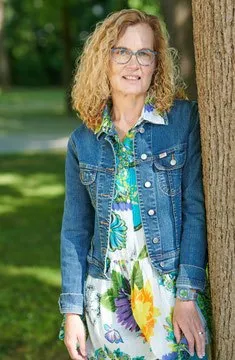 Eine Frau mit blonden lockigen Haaren steht draußen. Sie trägt eine Jeansjacke über einem Blumenkleid und eine Brille. Sie steht neben einem Baumstamm.