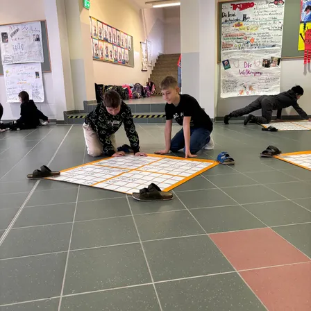 Zwei Jungen spielen ein Brettspiel auf einem gefliesten Boden, ein anderer Junge streckt sich in der Nähe. Andere Schüler sitzen und stehen herum, und es gibt Poster an der Wand.