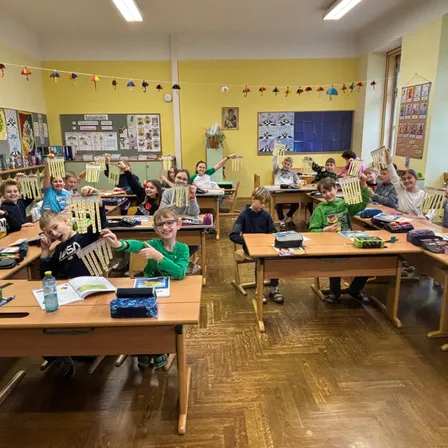 Klassenzimmer voller Schüler, die an Bastelarbeiten mit bunten Dekorationen arbeiten. Einige halten fertige Bastelarbeiten hoch, während andere an Tischen arbeiten.