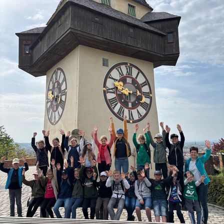 Eine Gruppe von Kindern posiert vor einem großen Uhrturm mit römischen Ziffern. Der Turm ist von Grün umgeben, und der Himmel ist teilweise bewölkt.