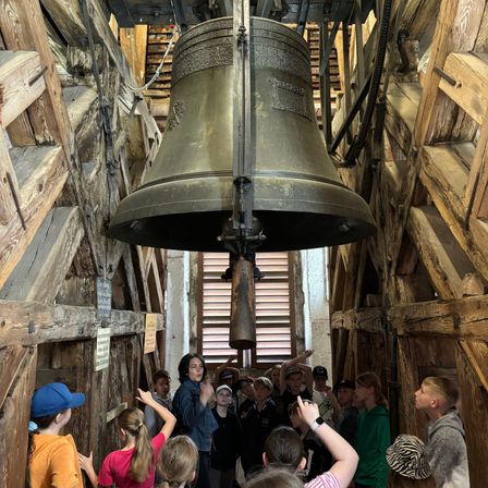 Eine Gruppe von Kindern und Erwachsenen schaut zu einer großen Glocke auf, die in einem hölzernen Raum von der Decke hängt.