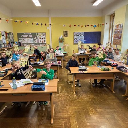 Klassenzimmer voller Schüler, die an Bastelarbeiten mit bunten Dekorationen arbeiten. Einige halten fertige Bastelarbeiten hoch, während andere an Tischen arbeiten.