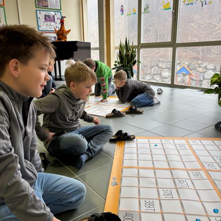 Eine Gruppe von Jungen spielt ein Spiel mit Kacheln auf dem Boden in einem Raum mit großen Fenstern und einem Klavier.