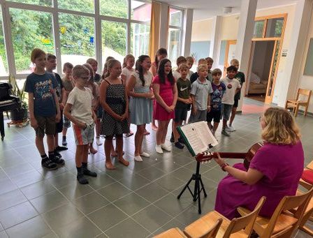 Eine Gruppe von Kindern singt, während eine Frau Gitarre spielt. Sie stehen in einer Reihe, mit einem Klavier im Hintergrund. Die Frau trägt ein lila Kleid.