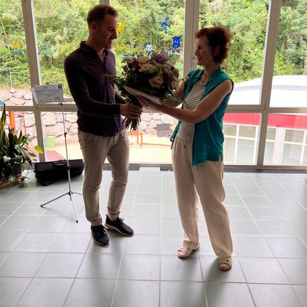 Ein Mann und eine Frau stehen in einem Raum und lächeln. Der Mann hält einen Blumenstrauß. Hinter ihnen sind Glasfenster mit Blick auf Bäume.