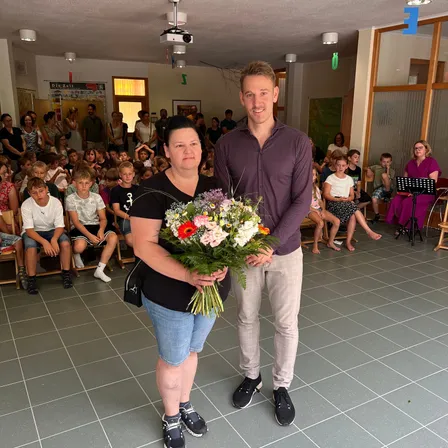 Ein Mann und eine Frau stehen in einem Raum mit Kindern auf Bänken, die einen Blumenstrauß halten. Der Mann lächelt.