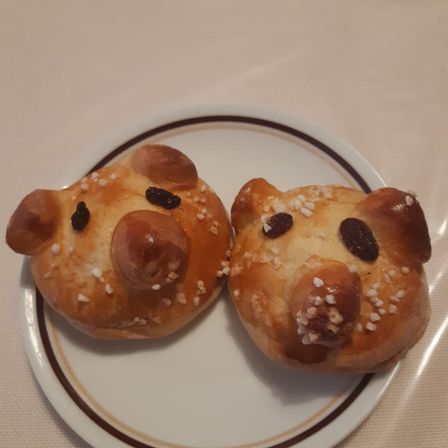 Zwei schweineförmige Gebäckstücke sind auf einem Teller dekoriert mit Rosinen und weißen Streuseln. Sie liegen auf einem weißen Tischtuch.