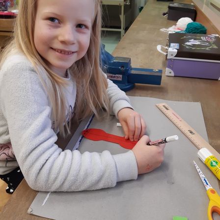Ein junges Mädchen mit blonden Haaren lächelt, während es auf einer grauen Oberfläche zeichnet, einen Marker und einen Lineal hält. Sie sitzt an einem Tisch mit Scheren und einer Box.