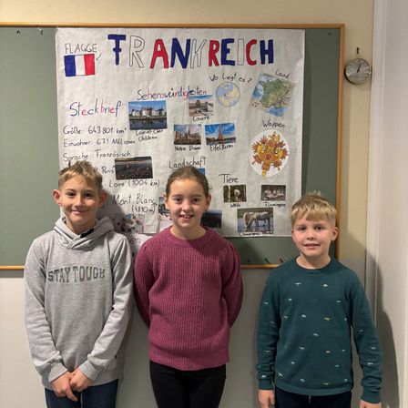 Drei Kinder stehen vor einem Poster über Frankreich. Das Poster enthält Text, Bilder französischer Sehenswürdigkeiten und eine Karte. Die Kinder tragen Freizeitkleidung.