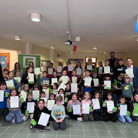 Eine Gruppe von Kindern und Erwachsenen steht und sitzt auf einem gefliesten Boden und hält Urkunden und grüne Boxen. Sie alle tragen Lächeln und posieren für ein Foto.