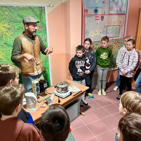 Ein Mann mit einer braunen Weste unterrichtet eine Gruppe von Kindern in einem Klassenzimmer mit einer Karte und einem Herd auf einem Tisch.