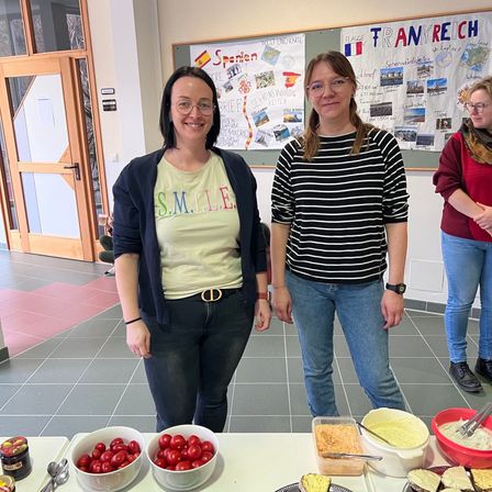 Drei Frauen stehen vor einem Tisch mit Essen. Eine trägt ein gelbes T-Shirt mit dem Wort 'S.M.I.L.E.' und eine andere ein gestreiftes T-Shirt. Sie stehen vor einem Bulletin Board mit einer Karte und Postern.