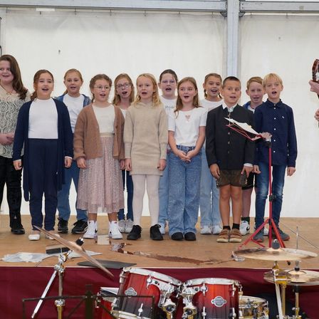 Eine Gruppe junger Kinder steht mit Musikinstrumenten und Mikrofonen auf der Bühne und nimmt wahrscheinlich an einer Aufführung teil.