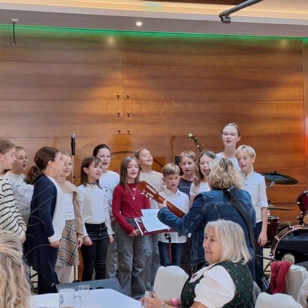 Eine Gruppe von Kindern und Erwachsenen, einschließlich einer Frau mit einer Gitarre, steht und singt vor einer Holzwand mit Mikrofonen und einem Schlagzeug.