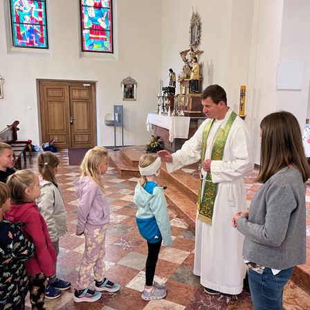 Ein Priester segnet Kinder in einer Kirche, umgeben von Altären und Buntglasfenstern.