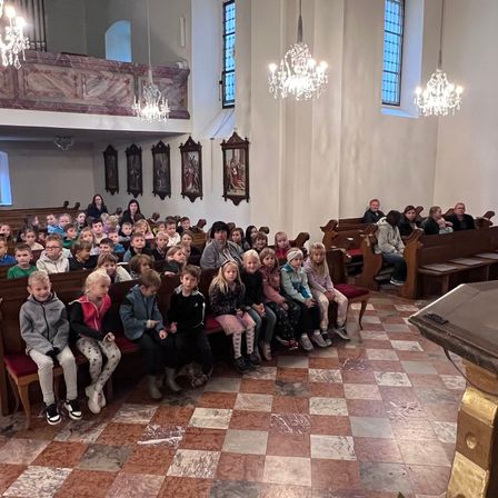 Eine Gruppe von Kindern sitzt auf einer Kirchenbank. Erwachsene sitzen hinter ihnen. Die Kirche hat Kronleuchter und Gemälde an der Wand.