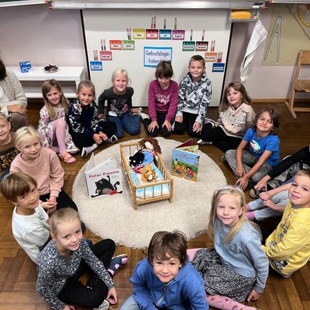 Eine Gruppe von Kindern sitzt im Kreis um ein kleines hölzerne Kinderbett, mit Spielzeug und Büchern darauf. Einige Kinder stehen in der Nähe des Bettes. Ein Kalender mit verschiedenen Monaten ist an der Wand befestigt. Ein weißer Tisch und ein Stuhl befinden sich auf der linken Seite.