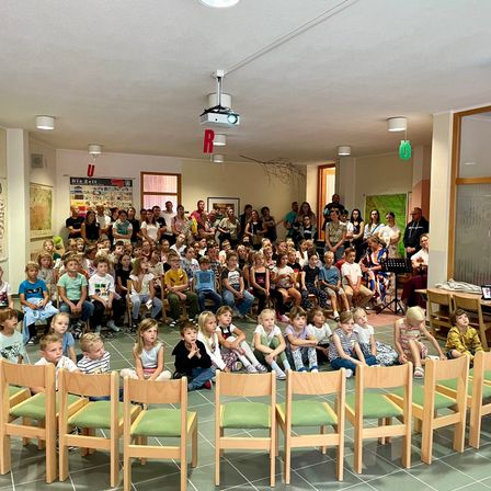 Eine große Menschenmenge aus Kindern und Erwachsenen sitzt in einem Raum zusammen. Einige sitzen auf Stühlen, andere stehen. Eine Projektorlampe befindet sich an der Decke.