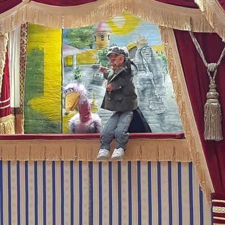 Ein Puppentheater-Auftritt mit einem Mann, der auf einem kleinen Vogel sitzt, beide in Kostümen. Der Hintergrund ist eine Gemälde mit einem Schloss und Bergen.