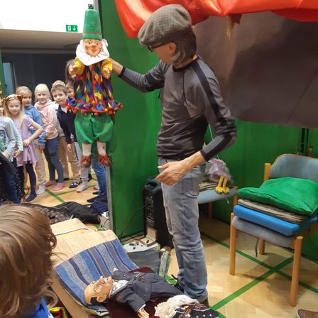 Ein Mann mit Mütze und Brille hält eine Puppe, während eine Gruppe von Kindern zusieht. Es gibt Kuscheltiere und einen Stuhl mit Kissen im Raum.