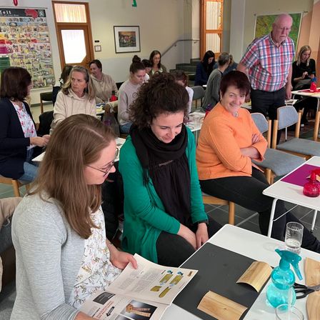 Eine Gruppe von Frauen sitzt in Stühlen, möglicherweise in einem Klassenzimmer. Eine Frau liest ein Buch, während eine andere sie ansieht. Vor ihnen stehen Tische mit verschiedenen Gegenständen.