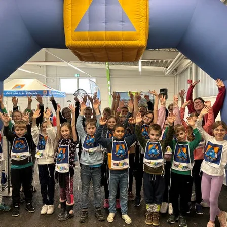 Eine Gruppe von Kindern posiert für ein Foto mit erhobenen Händen bei einer Indoor-Veranstaltung unter einem blauen Bogen. Sie tragen T-Shirts mit Designs, und eine Frau steht hinter ihnen.