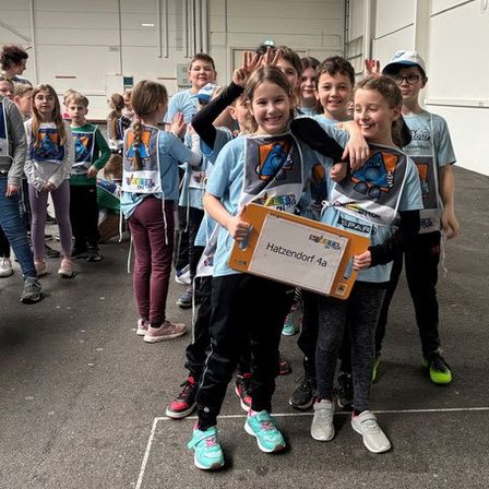 Eine Gruppe junger Schüler, die gleiche Hemden tragen, halten ein Schild und posieren für ein Foto. Sie stehen in einer Reihe, lächeln und scheinen sich in einer Schule zu befinden.