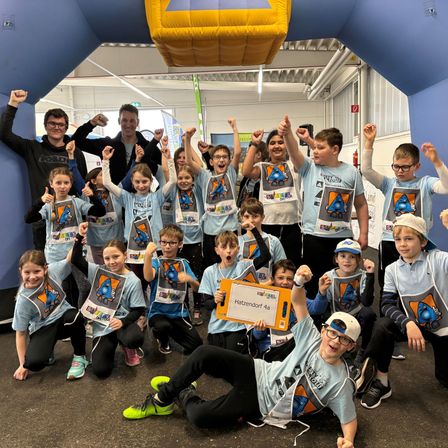 Gruppe von Kindern in passenden T-Shirts posiert für ein Foto, einer hält ein Schild, in einem blauen und gelben aufblasbaren Tunnel.