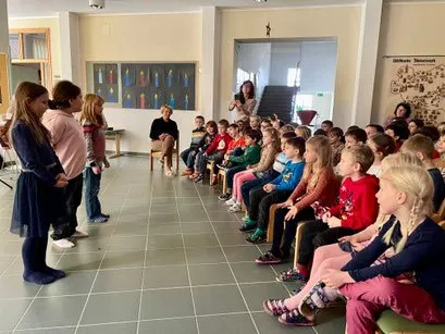 Mehrere Kinder sitzen auf Stühlen in einem Klassenzimmer, während eine Frau vor ihnen spricht. Andere Kinder stehen und lauschen, mit einer Treppe und einem Aushang im Hintergrund.