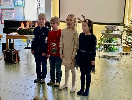Fünf Kinder stehen zusammen in einem Raum. Sie alle lächeln und scheinen zu singen. Ein Weihnachtsbaum und ein Klavier sind hinter ihnen.