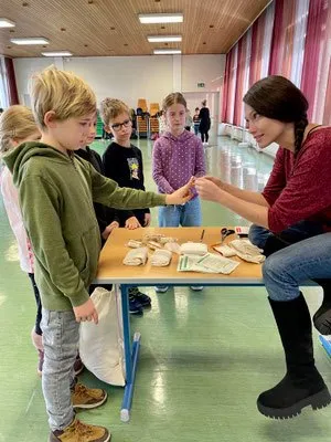 Eine Frau überreicht einem Jungen ein kleines Objekt an einem Tisch in einem Klassenzimmer, in dem Kinder stehen.