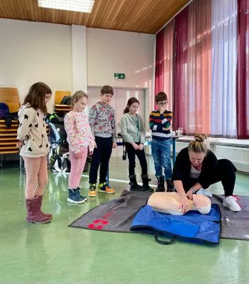 Eine Frau demonstriert CPR an einem Dummies, während mehrere Kinder in einem Klassenzimmer zusehen. Ein roter Vorhang und ein Heizkörper sind im Hintergrund.