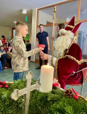 Ein Junge überreicht dem Weihnachtsmann ein Geschenk bei einer festlichen Veranstaltung. Der Weihnachtsmann sitzt in einem geschmückten Stuhl, hält einen Stab und eine brennende Kerze ist in der Nähe. Die Veranstaltung findet drinnen statt, mit einer Gruppe von Menschen im Hintergrund.
