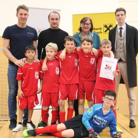 Eine Fußballmannschaft mit jungen Spielern in roten Trikots, begleitet von Trainern, steht vor einem Tor. Ein Junge in einem blauen Shirt hält ein Zertifikat.