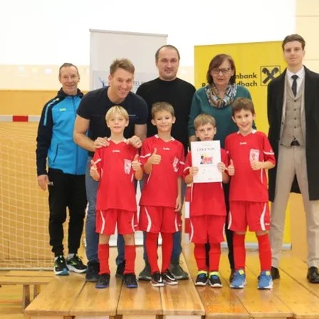 Fünf Kinder in roten Trikots stehen auf einer Bühne und halten Urkunden. Erwachsene stehen hinter ihnen. Sie befinden sich in einer Turnhalle mit gelber Wand und einem Tor.