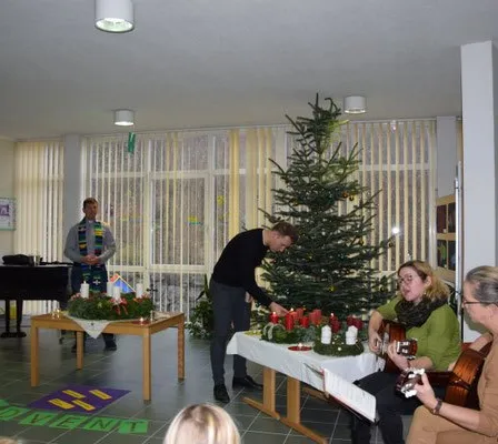 Eine Gruppe von Menschen versammelt sich in einem Raum mit einem Weihnachtsbaum, spielt Gitarren und singt. Ein Mann zündet Kerzen auf einem Tisch an. Ein anderer Mann steht neben einem Klavier.