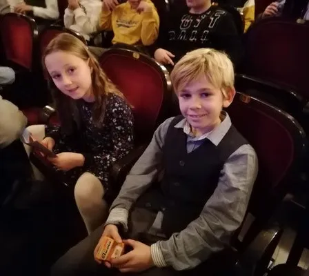 Ein Junge und ein Mädchen sitzen in Stühlen, wahrscheinlich in einem Theater, mit anderen Menschen im Hintergrund. Der Junge hält ein kleines Objekt, möglicherweise Süßigkeiten.