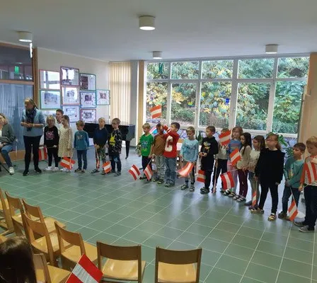 Eine Gruppe von Kindern und Erwachsenen steht in einem Raum und hält Flaggen. Eine Person steht mit einem Buch davor. Stühle sind um den Raum herum angeordnet. Es gibt Fenster mit Jalousien.