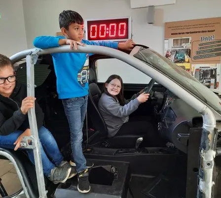 Drei Kinder stehen in einem Autogehäuse, ein Mädchen hält das Lenkrad. Ein Junge lehnt sich an die Tür, während ein anderer Junge im Inneren sitzt. Eine Digitaluhr ist an der Wand hinter ihnen.
