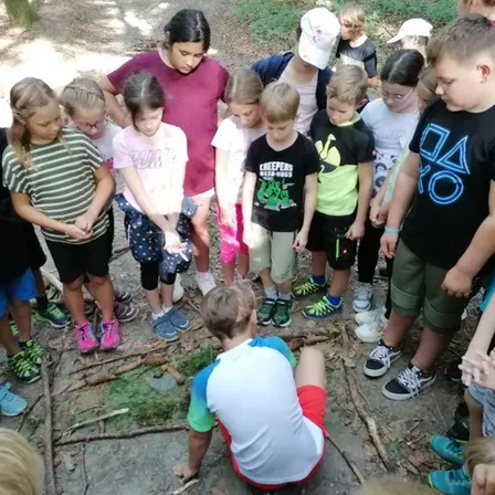 Eine Gruppe von Kindern und Erwachsenen ist im Freien und hat sich um einen kleinen Jungen versammelt, der auf dem Boden kniet. Sie tragen alle Freizeitkleidung.