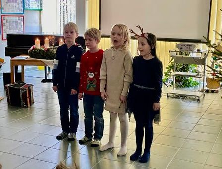 Fünf Kinder stehen zusammen in einem Raum. Sie alle lächeln und scheinen zu singen. Ein Weihnachtsbaum und ein Klavier sind hinter ihnen.