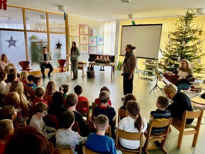 Eine Gruppe von Menschen, darunter Kinder, sitzen in einem Raum mit einem Weihnachtsbaum und lauschen einer Präsentation vor einer Projektionsleinwand. Zwei Personen stehen und sprechen.