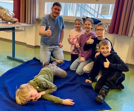 Ein Mann und fünf Kinder posieren für ein Foto. Ein Kind liegt auf einer blauen Decke, während die anderen sitzen. Sie alle zeigen Daumen hoch. Ein Tisch mit einem Korb ist im Hintergrund.