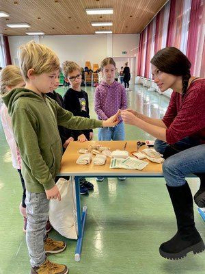 Eine Frau überreicht einem Jungen ein kleines Objekt an einem Tisch in einem Klassenzimmer, in dem Kinder stehen.