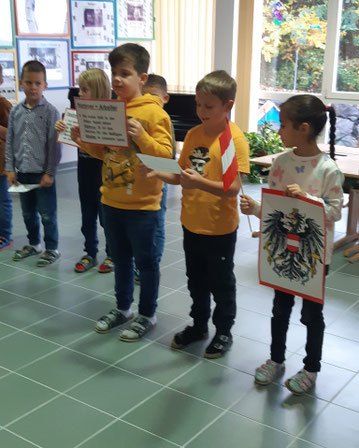 Eine Gruppe junger Kinder steht in einem Klassenzimmer und hält eine Flagge und ein Zertifikat. Einige tragen Turnschuhe, andere haben Schuhe an. Sie posieren für ein Foto.