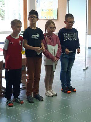 Vier Kinder stehen in einem Raum. Drei sind Jungen, eines ist ein Mädchen. Der Junge in der Mitte trägt eine Brille und hält ein Papier. Der Junge auf der rechten Seite hat orange Schuhe.