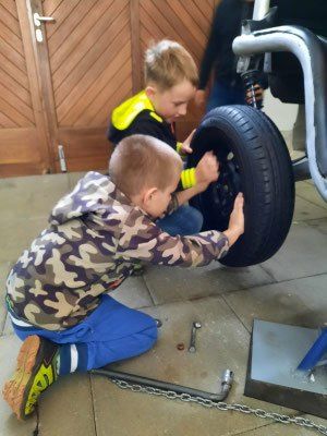 Zwei Jungen arbeiten an einer Autoreifen. Ein Junge hält einen Schraubenschlüssel, während der andere das Rad hält. Sie sind in einem Raum mit einer Holztür.