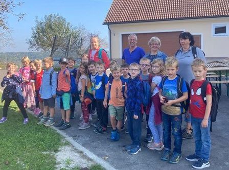 Eine Gruppe von Kindern mit Lehrern steht an einem sonnigen Tag vor einem Haus. Sie tragen alle Rucksäcke und Turnschuhe.