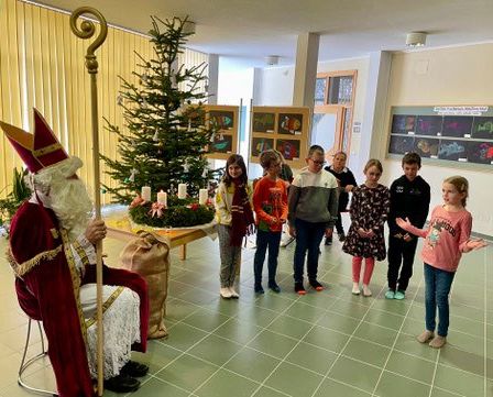 Der Weihnachtsmann in rot und weiß sitzt in einem Stuhl, umgeben von Kindern, die in einem Klassenzimmer singen.